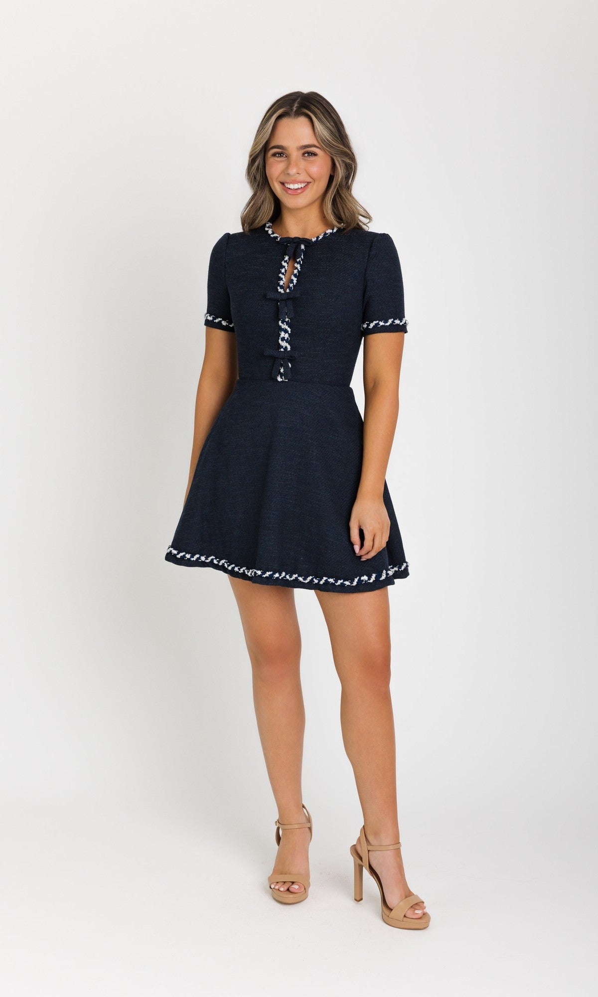 Smiling and facing forward, a woman poses against a white background in the Ava Presley 42927 Short Homecoming Dress in navy, featuring decorative trim and paired with nude heels—perfect for any homecoming event.